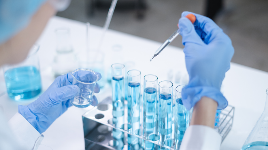 A képen egy laboratóriumi munkatárs látható, aki kék folyadékkal teli kémcsöveket és egy kémcsőbe csepegtető pipettát használ. Az asztalon különböző üvegek és kémcsövek vannak.