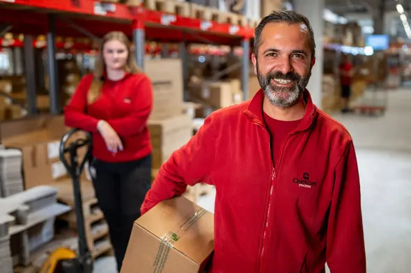 Két dolgozó látható egy raktárban. Az elülső férfi mosolyogva tart egy dobozt, míg a háttérben egy nő áll, aki szintén egy vörös pulóvert visel. A raktárban dobozok és árut mozgató eszközök találhatók.