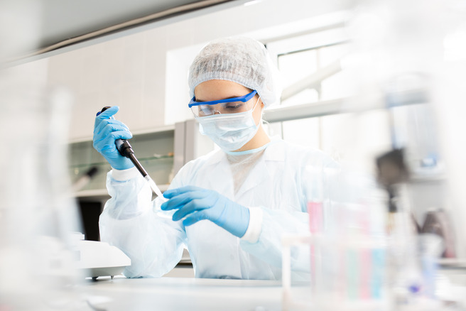 Egy laboratóriumban dolgozó kutató, aki pipettát használ. A nő védőfelszerelést visel, beleértve az arcvédőt és kesztyűt. A háttérben laboratóriumi felszerelések láthatók.