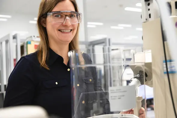 Laboratóriumban dolgozó nő, védőszemüvegben, mosolyogva áll egy berendezés mellett. A háttérben laboratóriumi felszerelések és munkaállomások láthatók.
