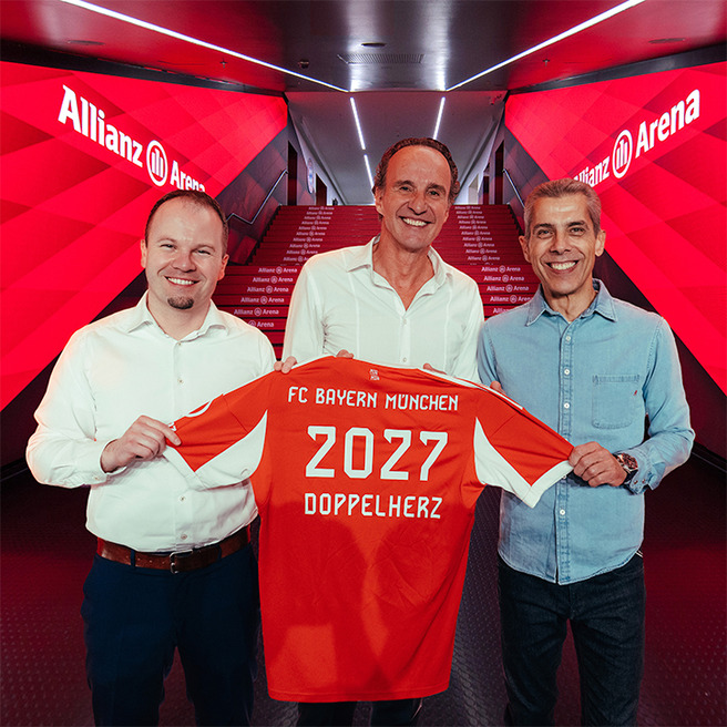 Három férfi áll egy színes háttér előtt, és bemutat egy piros focimezt. A mez hátán "FC Bayern München", "2027" és "DOPPELHERZ" feliratok láthatók. Az esemény az Allianz Arenában zajlik. Az arcuk boldog kifejezését mutatja az együttműködés örömére.