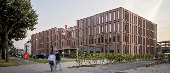 Modern, téglaburkolatú irodaház látható, amely az utcán áll. A közelben néhány autó áll, emberek sétálnak az épület előtt. Az épület mellett zöld növényzet található, a háttérben fák állnak.