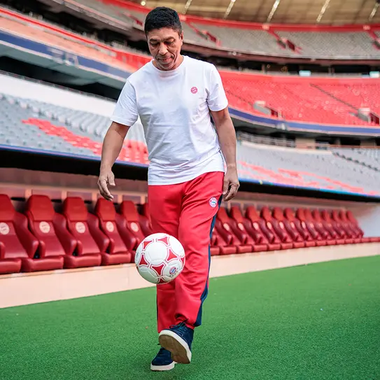 Egy férfi, fehér pólóban és piros sportnadrágban, futballlabdát rugdos egy üres stadionban. A háttérben piros székek és a lelátó látható. Az férfi mosolyogva játszik, a zöld fűn halad előre.