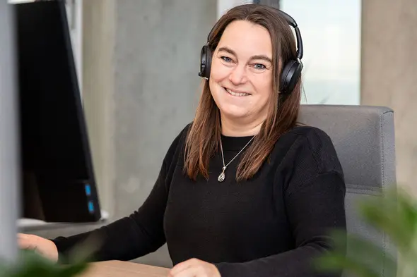 Egy nő mosolyogva ül egy irodai széken, fején fülhallgatóval. Előtte egy számítógép található, és barátságos, segítőkész légkör érződik a képen.