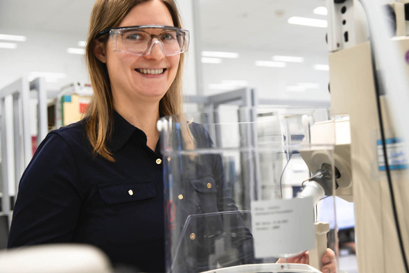 Laboratóriumban dolgozó nő, védőszemüvegben, mosolyogva áll egy berendezés mellett. A háttérben laboratóriumi felszerelések és munkaállomások láthatók.